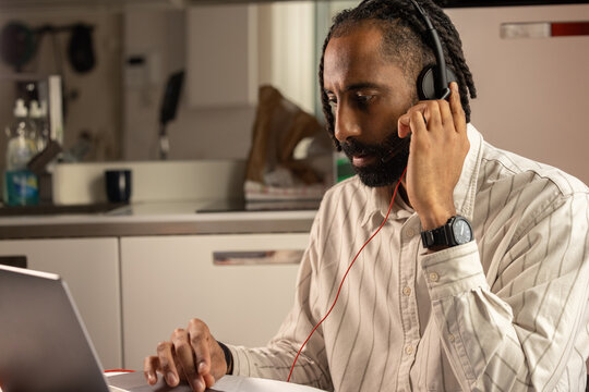 Portrait d'un jeune homme noir de 30 ans qui travaille &agrave; la maison en t&eacute;l&eacute;travail. Il est assis devant un ordinateur portable et il a un casque audio sur les oreilles