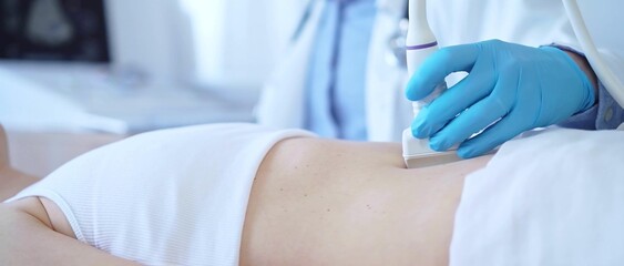 Fototapeta premium Doctor man wearing a blue medical gloves using an ultrasound equipment on a female patient lying down in clinic cabinet, close up view. Medicine concept
