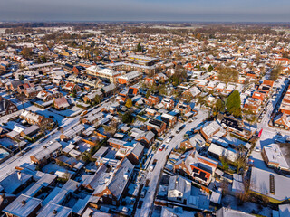 Obraz premium Aerial view of a dense residential neighborhood in a Dutch town after snowfall. A winter landscape featuring rows of red brick houses with snow covered roofs under a clear blue morning sky.