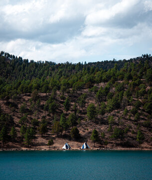 Two white tipis on the rocky shore of a blue mountain lake with a dense pine forest hillside under a cloudy sky