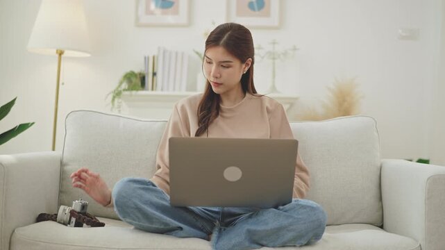 Happy Asian Vlogger or Creator Woman sitting on a cozy sofa. She smiling excitedly while holding camera in one hand and using a laptop computer search for hotels or tourist attractions.