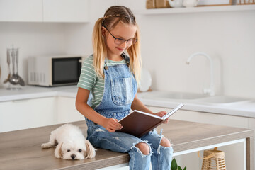 Fototapeta premium Cute little happy girl with Maltese dog sitting on table and reading book in kitchen