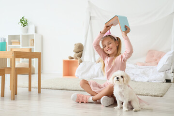 Fototapeta premium Cute little happy girl in princess crown with Maltese dog reading book on floor at home