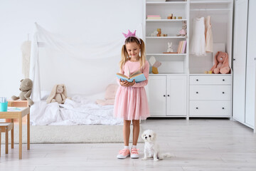 Fototapeta premium Cute little happy girl in princess crown with Maltese dog reading book at home