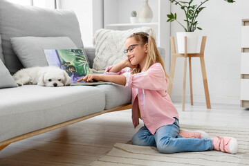 Fototapeta premium Cute little happy girl with Maltese dog reading book on floor near sofa at home