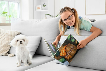 Fototapeta premium Cute little happy girl with Maltese dog reading book on sofa at home