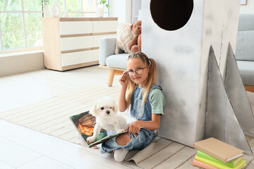 Fototapeta premium Cute little happy girl with Maltese dog and books sitting on floor near cardboard space rocket at home