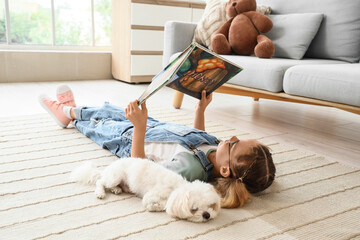 Fototapeta premium Cute little happy girl with Maltese dog reading book on floor at home