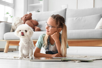 Fototapeta premium Cute little happy girl with Maltese dog reading book on floor at home