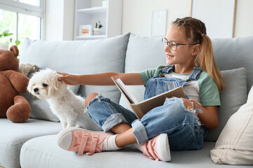 Fototapeta premium Cute little happy girl with Maltese dog reading book on sofa at home