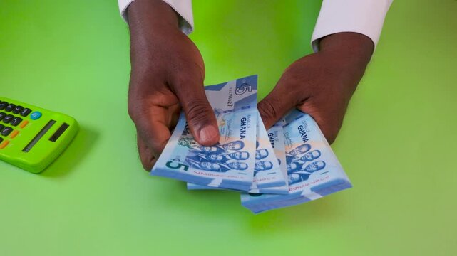 A high-angle shot shows a man's hands counting blue Ghana 5 Cedi notes over a bright green desk. A matching lime green calculator sits nearby, emphasizing themes of accounting and finance.