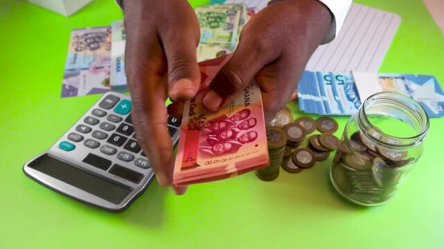 A person counts Ghanaian Cedi notes and organizes coins into a glass jar. Using a calculator and notebook, they demonstrate careful financial budgeting and the importance of personal savings.