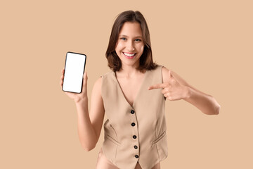 Naklejka premium Beautiful young happy woman in stylish vest pointing at mobile phone on beige background