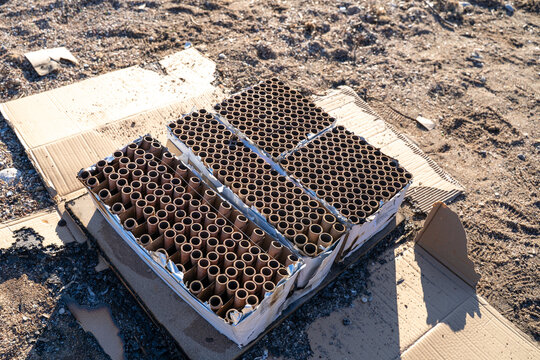 Spent fireworks cake boxes with empty cardboard launch tubes sitting on flattened cardboard on sandy ground, aftermath of pyrotechnic celebration with ash and debris in bright sunlight