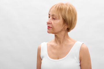 Mature woman with short blonde hair wearing white sleeveless top looks aside on plain background....
