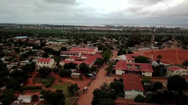 Aerial view of a suburban neighborhood in Maputo, Mozambique, with red‑roofed houses, dirt roads, scattered trees, and the distant city skyline and bay under an overcast sky.