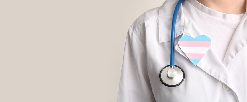 Doctor with paper heart in colors of transgender flag on light background with space for text, closeup