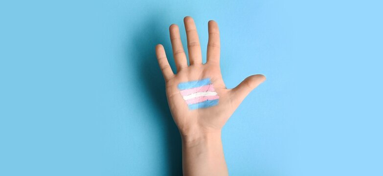 Hand with painted transgender flag on light blue background, top view
