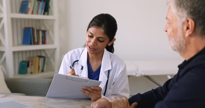 Happy young Indian female doctor in white coat talking to elder patient and taking notes at workplace, keeping records, getting happy, smiling, giving handshake to senior man, finishing meeting