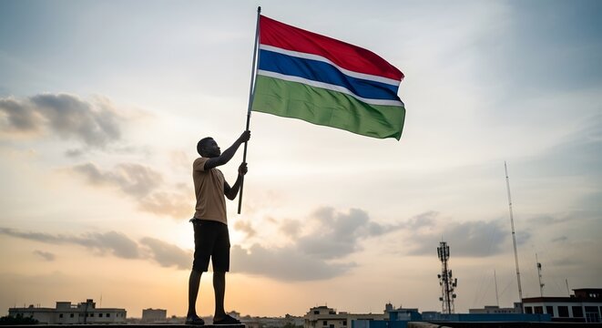Silhouetted figure proudly raising the Gambian flag against a dramatic sunset sky, symbolizing national identity.