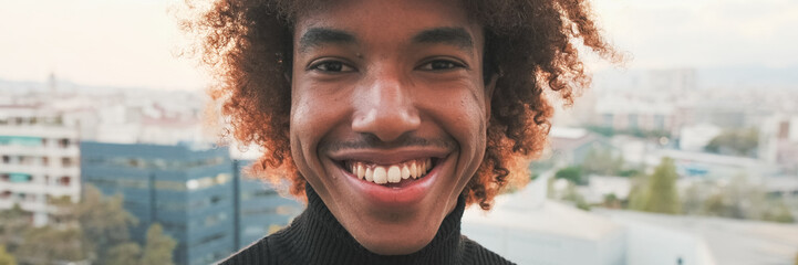 Fototapeta premium Close-up of young smiling guy posing at the camera while standing on the balcony modern city on the background. Panoramic