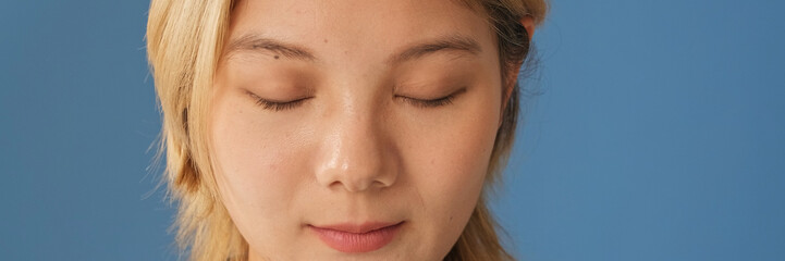 Fototapeta premium Close up of young woman dressed in denim shirt stands with her eyes closed isolated on blue background in studio. Panoramic
