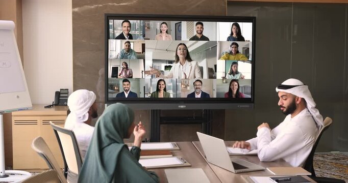 Global business partners meeting on hybrid conference. Team of Arabic business people sitting at large monitor with collage of head shots, talking to colleagues on video call