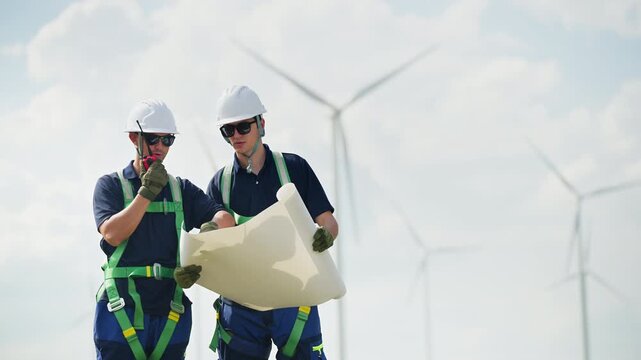 Engineers wearing safety helmets inspecting a wind turbine at a wind farm holding a blueprint while working. renewable energy sustainable technology wind power maintenance environmental engineering.