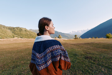 Naklejka premium Woman in patterned sweater stands in a mountain landscape, overlooking a wide meadow and clear sky. Outdoor portrait conveys calm nature scene, travel and peaceful solitude.