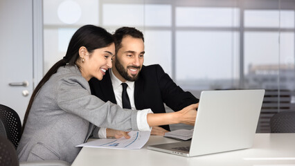 Fototapeta premium Smiling business colleagues reviewing data on laptop in office, analyzing reports on screen, discussing corporate strategy, financial performance and online project result. Collaboration, teamwork