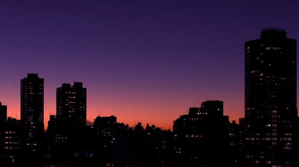 Fototapeta premium Silhouetted cityscape at dusk with glowing windows against a vibrant purple and orange sky