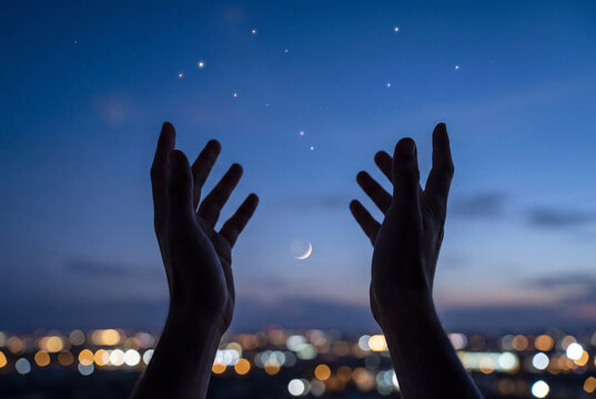 Silhouette of hands reaching up to the stars and crescent moon in the night sky over a city