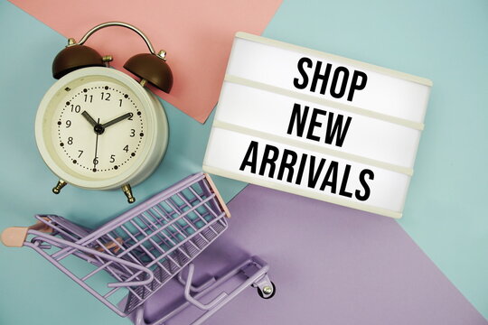 Shop New Arrivals text in lightbox with trolley shopping cart and alarm clock flat lay on multicolor pastel background