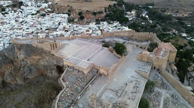Cinematic drone orbit of the Lindos Acropolis during blue hour. The shot reveals the traditional white village and the scenic harbor at dusk. Iconic ancient landmark and Mediterranean architecture