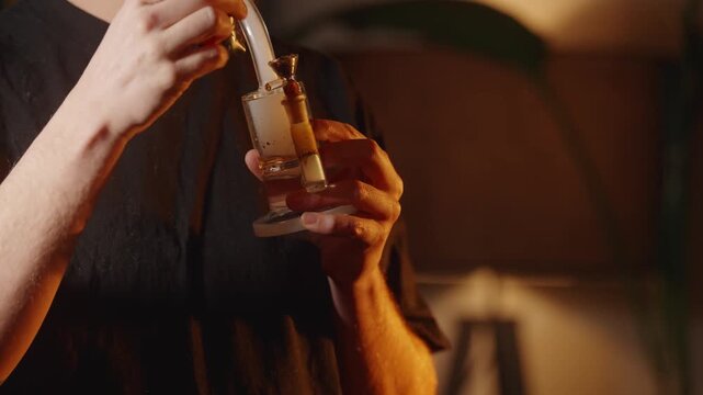Man using glass bong to smoke cannabis indoors with warm lighting.