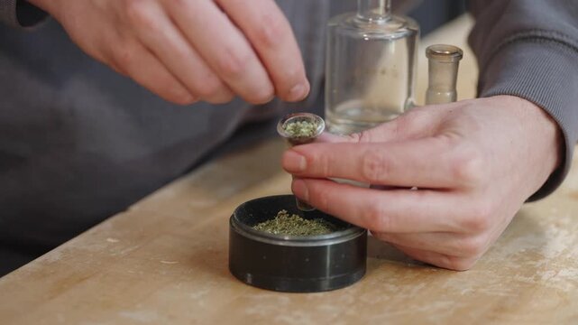 Hands preparing ground cannabis using metal grinder before smoking with glass bong.