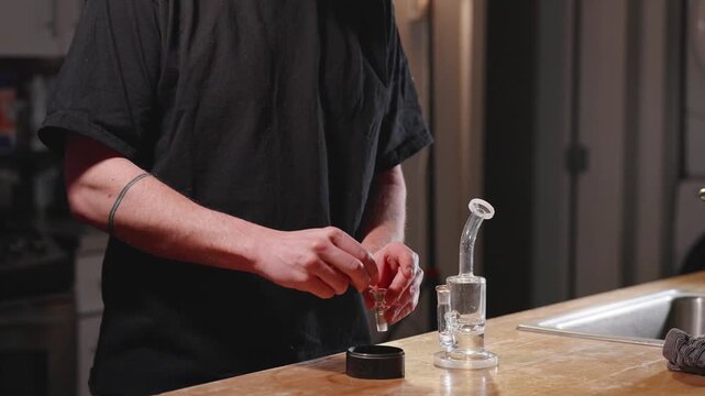 Person preparing cannabis with grinder beside glass bong on kitchen counter.