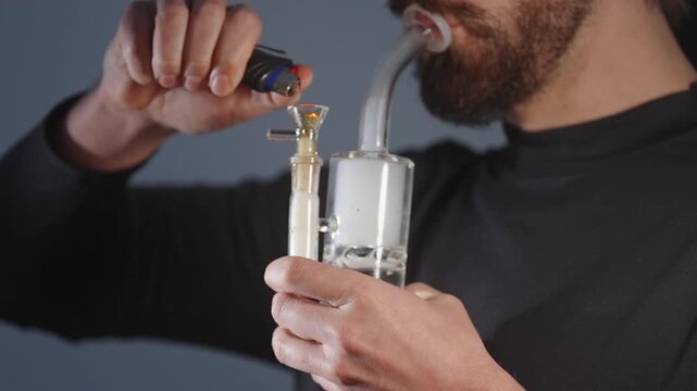Close view of man lighting a glass bong with lighter while preparing to smoke cannabis.