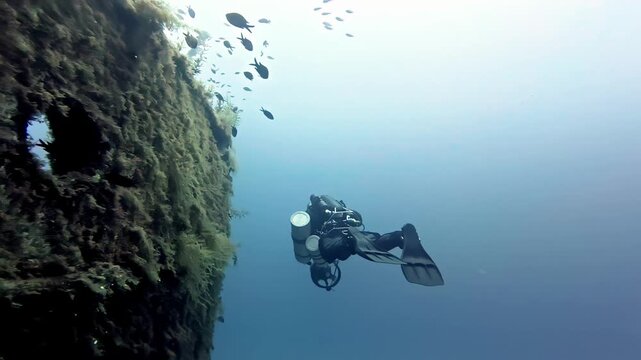 Professional technical scuba diver exploring deep rustic shipwreck hull in clear blue Red Sea waters during summer morning with schooling tropical fish and high angle wide shot cinematic lighting