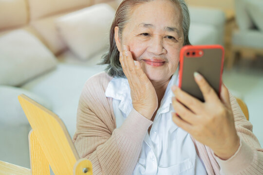 Happy older Asian woman touching face skin while looking her face on smartphone