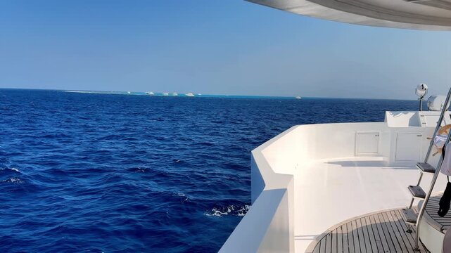 A pristine white luxury yacht deck glides through deep sapphire Red Sea waters under a clear summer sky with distant coral reefs and white boats visible from a high-angle panoramic perspective