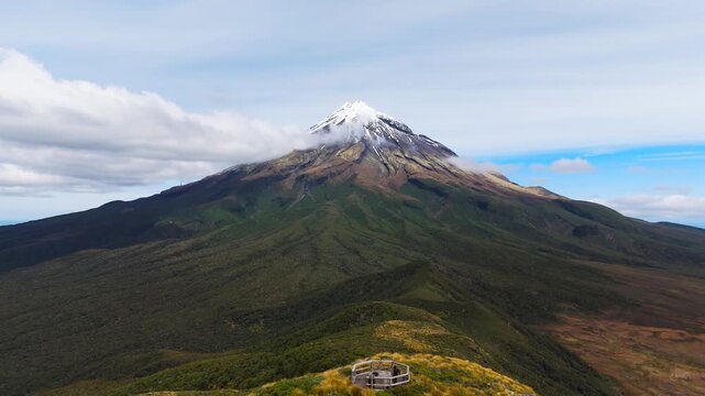 Majestic Mount Taranaki in New Zealand, the snow-capped volcanic peak, forested slopes and a scenic viewpoint beneath drifting cloud, cinematic aerial dolly view