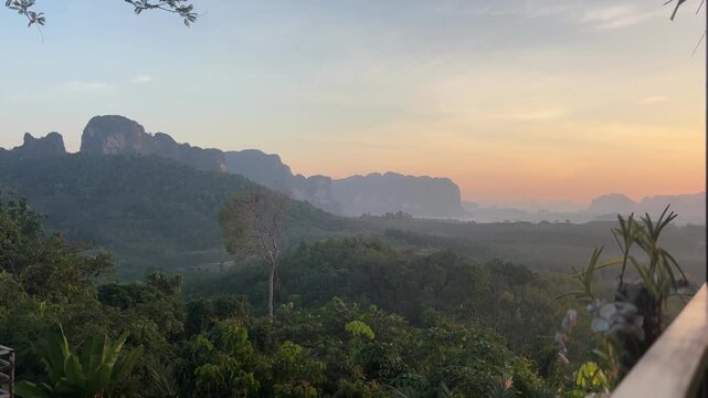 Sunrise light spreading across forest valley and distant limestone hills at Din Daeng Doi in Krabi Thailand with warm sunlight, soft haze, and calm tropical landscape