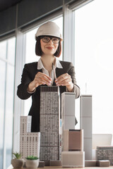 Fototapeta premium A smiling female architect wearing a hard hat and glasses uses a compass to measure architectural models of skyscrapers in an office setting