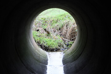 Fototapeta premium View through a round concrete tunnel carrying a stream beneath a road