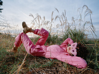 Naklejka premium Outdoor fashion in a sunny meadow features a woman in a pink jumpsuit lying among tall grasses, relaxed pose, playful mood, and natural light emphasizing texture and color.