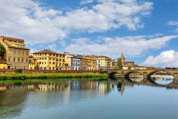 Fototapeta premium Santa Trinita Holy Trinity bridge gracefully crosses the Arno River in Florence, highlighting the beauty of medieval architecture against a backdrop of lively buildings and a blue sky.