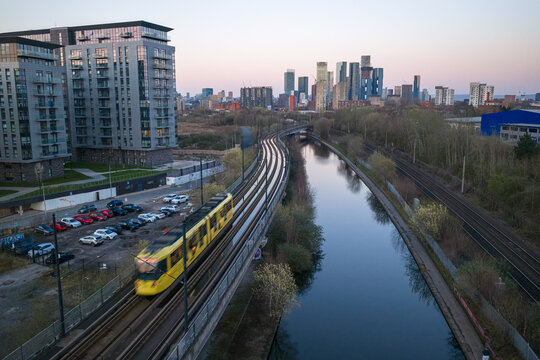 Aerial image of a moving yellow Metrolink tram passing Pomona Wharf toward the Deansgate skyscrapers in Manchester, England, UK.
