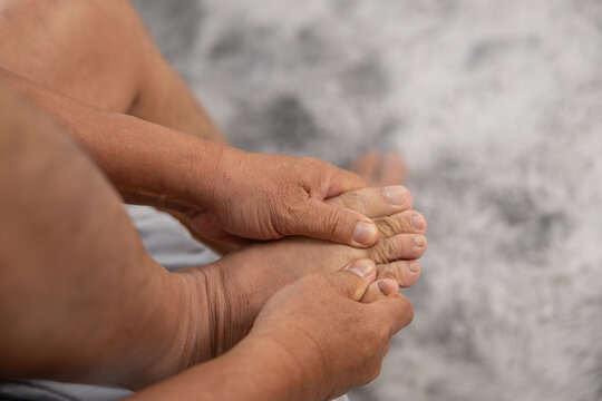 Senior adult hands gently massaging tired bare toes experiencing severe foot pain and uncomfortable muscle ache while sitting on soft gray carpet floor feeling physically exhausted