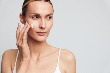 Fototapeta premium Young woman applying face cream on clean skin, skincare routine, natural beauty, soft lighting, white background, focused expression, casual wear, healthy lifestyle concept